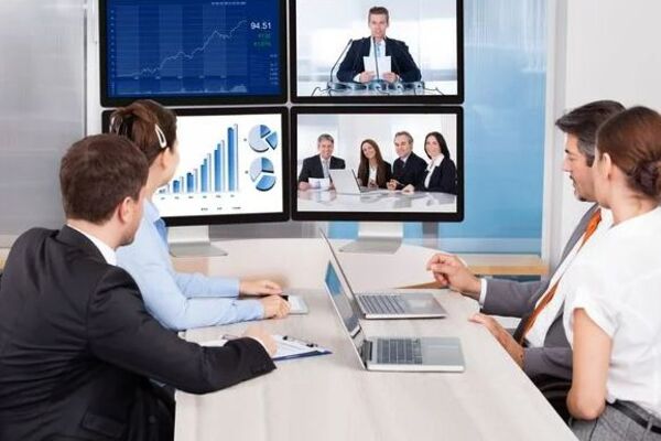 Personen an einem Tisch sitzend schauen auf mehrere Bildschirme für eine Videokonferenz.