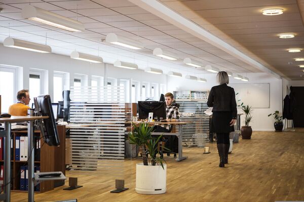 Büro mit mehreren Schreibtischen und Personen an einem langen Gang.